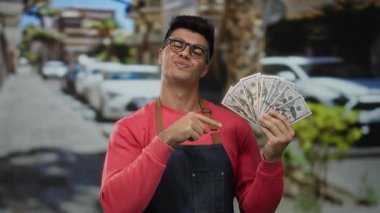 Kentsel bir sokakta Amerikan dolarlarını elinde tutan adam, sıradan giyinmiş, bulanık şehir arka planında park halindeki arabalarla güvenli bir şekilde para gösteriyor..