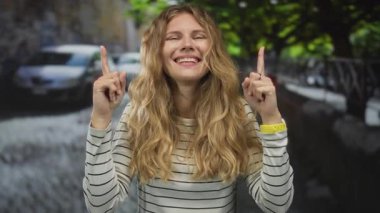 Sokakları gösteren parmakları olan bir kadın, açık hava ortamında gülümseyen genç bir sarışın gibi görünüyor..