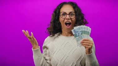 Brezilya banknotları tutan orta yaşlı bir kadın canlı mor arka plana karşı şaşkın bir ifadeyle.