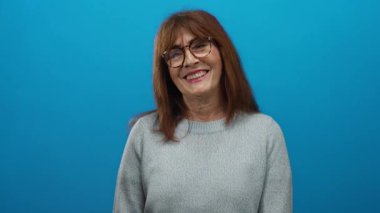 İzole edilmiş mavi bir duvara sıcak bir şekilde gülümseyen, gözlüklü, rahat bir kazak giyen, sıcaklığını ve güvenini tarafsız bir ortama aktaran İspanyol son sınıf öğrencisi bir kadın..