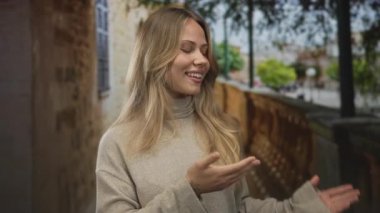 Sarı saçlı ve gülümseyen yüzlü bir kadın avuçlarını dar bir sokakta eski bir duvarın ve sağlam bir korkuluğun yanında sunuyor..