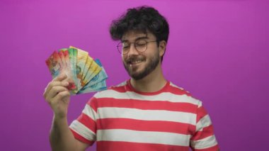 Çizgili tişörtlü, sakallı, parlak pembe arkaplanlı İsviçre frangına gülümseyen ve işaret eden genç İspanyol adam coşku ve gururla renkli para birimi notları gösteriyor..