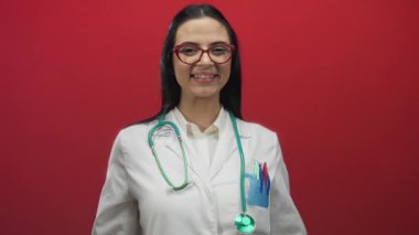 Steteskoplu ve gözlüklü İspanyol kadın doktor canlı kırmızı duvarlara karşı güvenle gülümsüyor. Sağlık sektöründe profesyonelliği gösteriyor..
