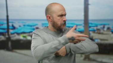 Sakallı kel adam, bulutlu bir günde arka planda mavi şemsiyelerle kumlu deniz kenarına halat çekerek oyuncu ve yaratıcı havayı yansıtıyor..