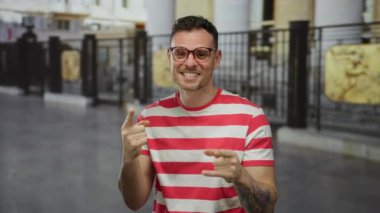 Çizgili tişörtlü genç adam başparmağını kaldırıyor ve kent geçmişiyle gülümsüyor. Şehir sokaklarında pozitif ve kendine güvenini gösteriyor..