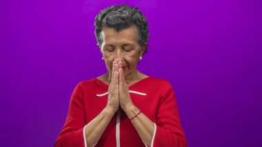 Gözleri kapalı dua eden, kırmızı elbise giyen, mor arka plana karşı duran, gri saçlı son sınıf öğrencisi, dinginliği tasvir eden, ruhani, huzurlu, sakin, yaşlı, odaklanmış bir kadın..