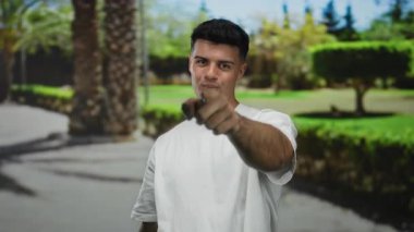 Genç adam, canlı yeşil ağaçlar ve güneş ışığının gölgelerinin olduğu açık hava parkına güveni ile işaret ederek doğal bir ortamda pozitif ve çekici bir tavır sergiliyor..