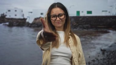 Kadın deniz kenarı binasının önünde el kamerasına uzanır, gözlük ve günlük gömlekle gülümser; dostça selamlaşır.