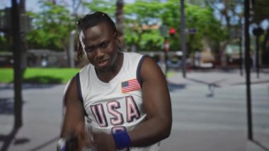 ABD forması giyen genç adam kameraya doğru bakıyor ve ağaçlar ve trafik ışığı görünen güneşli bir caddede yavaşça yürüyor. Kararlılık gururu..