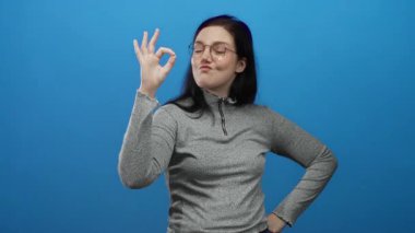 Esmer saçlı bir kadın, gri bir süveter ve gözlüklü, parlak mavi bir arka plana karşı güvenle gülümseyen hoş bir jest yapıyor..