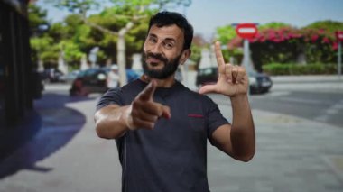 Sakallı İspanyol bir adam şakacı ezik hareketler yapıyor ve ağaçlar ve park edilmiş arabalarla dolu neşeli bir şehir caddesini işaret ediyor. Espri ve alaycılık yapıyor..