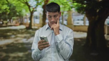 Açık mavi tişörtlü adam elinde akıllı telefon ile yeşil parktaki çeneye dokunuyor ve güneş ışığı ağaçların arasından süzülüyor..
