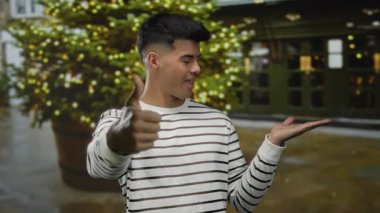 Genç adam el hareketleriyle şehir sokaklarında, çizgili gömlek giyiyor, arka planda Noel ışıkları, neşeli tatil ruhunu taşıyor..