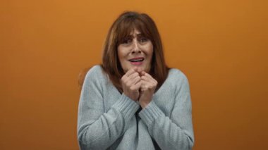 Üst düzey İspanyol kadın kazak giyiyor turuncu bir duvara karşı korkmuş görünüyor, canlı ve izole bir ortamda bir duygu anı yakalıyor..