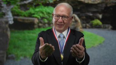 Takım elbiseli kıdemli bir adam, elleri parktaki yeri işaret ediyor, madalya ve gözlük takıyor, açık havada yeşillikli bir yerde ödül jesti yapıyor..