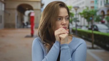 Bulanık arabaların ve sokak lambalarının arasında ellerini sıkan gergin ifadeye sahip genç beyaz kadın; endişe.