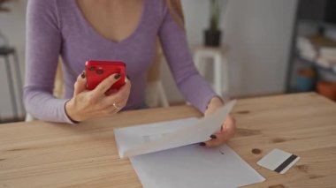 Oturma odasında elinde akıllı telefon ve belge olan bir kadın, etrafı kredi kartı ve evrak işleriyle çevrili olan verileri analiz ederken, evde mali planlama yapıldığını öne sürüyor..