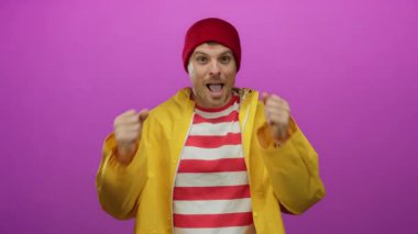 Sarı yağmurluk ve kırmızı bereyle neşeli bir ifadeyle kutlayan adam canlı pembe arka plana karşı gençlik heyecanı ve pozitifliği somutlaştırıyor..