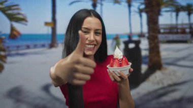 Sahilde dondurmadan hoşlanan bir kadın, arka planda palmiye ağaçları olan kırmızı bir tişörtle güneşli bir gezinti yaparken baş parmağını kaldırıyor..