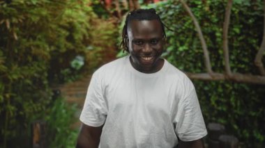 Elleri havada bir adam, avuçları önde, yeşil parkta beyaz tişört giymiş ve gülümsüyor, kameraya bakıyor..