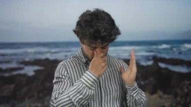 Okyanusun kenarında dalgalar bulutlu bir atmosferde kayalara çarparken kötü kokudan dolayı deniz kenarına burnunu sokan sakallı genç bir adam..