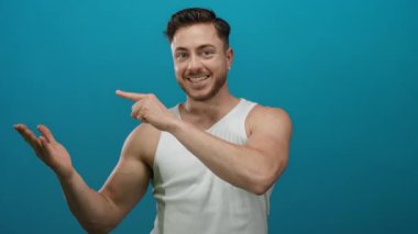 Beyaz atletli sakallı genç adam gülümsüyor ve açık bir eli işaret ediyor. Mavi arka plana karşı. Pozitif ve davetkar bir sunum yapıyor..