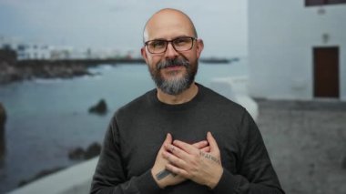 Deniz kenarındaki gezinti alanının önünde elleri dövmeli gülümseyen gözlüklü, süveterli bulanık plajlı ve arka planda binalar olan bir adam..