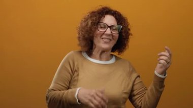 Orta yaşlı İspanyol kadın, neşeli bir ifade ve kıvırcık saçlarla yaratıcılık ve neşeyi kucaklayan, izole turuncu bir arka planda gitar çalıyormuş gibi yapıyor..