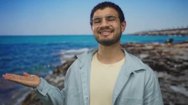 Genç İspanyol adam gülümseyen sol palmiyesini uzatarak turkuaz denizi ve güneşli sahilde kayalık kıyı şeridini sunar; dinginlik.