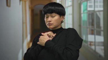 Kapalı bir otel koridorunda elleri sımsıkı bağlanmış, siyah süveter giyen, iyi aydınlatılmış bir şehir ortamında gergin bir anı yakalayan bir kadın..