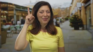 Orta yaşlı, sarı tişörtlü İspanyol kadın sıcak bir şekilde gülümsüyor ve şehir caddesinde parmakla işaret ediyor. İyimserlik..