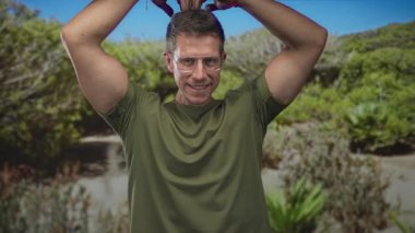 Elleri başının üstünde kalp oluşturan bir adam, gülümseyen ve bir orman yolunda gözlük ayarlayan zeytin yeşili gömlek ve bilezik giyen kaygısız neşe.