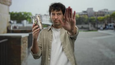 Açık havada para dolu bir kumbarayı elinde tutan bir adam, durması için el işareti yapan, kentsel bir ortamda finans ve ihtiyatı vurgulayan bir adam..