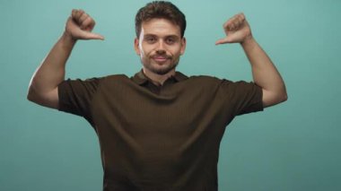 Stüdyoda gülümserken başparmaklarını göğsüne gösteren adam, görünür eller ve ön kollar deniz mavisi arkaplanına karşı; gururlu güven karizması.