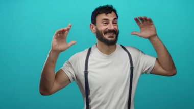 Sakallı İspanyol bir adam, pantolon askısı takıyor, neşeli ve neşeli bir enerji saçan, parlak mavi bir arka plana karşı şakacı hareketler yapıyor..