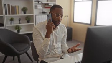 Beyaz gömlekli bir adam telefonla başa baş konuşuyor ve ofis masasında gözlük takıyor..