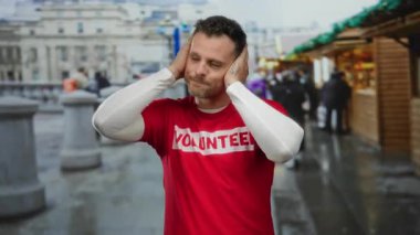 Kırmızı gönüllü gömleği giyen genç bir adam kalabalık bir şehir caddesinde dikilirken kulaklarını kapatır, canlı bir şehir atmosferi olduğunu gösterir..