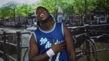 Adam gülümsüyor ve mavi basketbol forması içinde kameraya doğru işaret ediyor. Kanal korkulukları, Amsterdam şehri sahnesi, neşe yoldaşlığı..