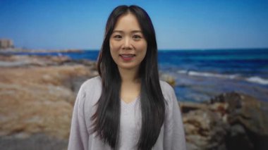 Deniz kenarındaki kumsalda gülümseyen genç Çinli kadın berrak mavi gökyüzü ve arka planda sakin okyanus dalgaları açık havada sükunetini sergiliyor.