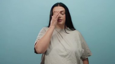 Geniş cüsseli bir kadın, izole edilmiş mavi bir arkaplanda dikiliyor ve yüzünü sıradan bir şekilde kapatıyor.