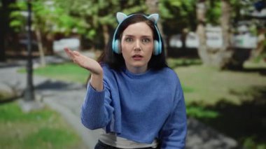 Kulaklık takan ve yeşil park ortamında özellikle el kol hareketi yapan bir kadın dışarıda duygusal bir ifade sergiliyor..