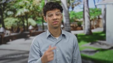 Gözlüklü ve açık mavi gömlekli bir adam, yeşil parkta bulanık banklar ve ağaçlarla parmak yanağa duruyor..