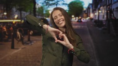 Gün batımında gülümseyen ve yan bakan, kaldırımlı bir şehir caddesinde elleri ile bir kalp oluşturan genç İspanyol kadın; sevgi sıcaklığı.