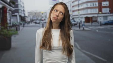 Uzun kahverengi saçlı, beyaz bluzlu bir kadın, binalar ve kalın saçlarla bir şehir caddesinde somurtuyor ve yuvarlanan gözler;.