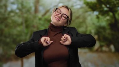 Parkta gözlük ve balıkçı yaka giyen yeşil ağaçlarla çevrili bir kadın güvenli bir şekilde gülümsüyor. Huzurlu bir doğal ortam yaratıyor.