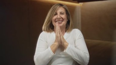 Kahverengi panellere karşı sıcak ışık altında, elleri göğsünde birbirine bastırılmış, orta yaşlı beyaz bir kadın..