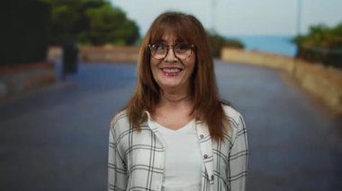 Üst düzey İspanyol kadın gülümsüyor ve açık bir şehir caddesinde kareli gömlek ve gözlük takıyor, arkasında ağaçlar ve okyanus var, özgüvenini ifade ediyor..