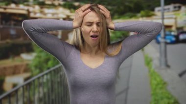 Stresli bir kadın, şehir sokağında elleri başının üstünde, şehir manzarasının ortasında hayal kırıklığını ifade ediyor, endişeli bir tavırla günlük giysiler giyiyor..