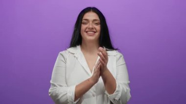 İzole edilmiş mor arka planda, beyaz gömlek giyen, kendine güveni ve pozitifliğiyle mutlu bir şekilde alkışlayan kadın..