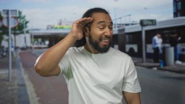 Otobüs durağında, beyaz tişört giymiş, otobüs ve kaldırım kenarında el pençe divan duran bir adam..